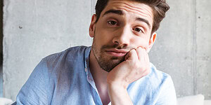 Thoughtful young man with a friendly expression, sitting indoors with a concrete wall background, illustrating themes of psychology, mental health, and emotional well-being.