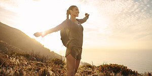 A woman enjoying outdoor nature hike, spreading arms joyfully against a scenic coastal background at sunrise.